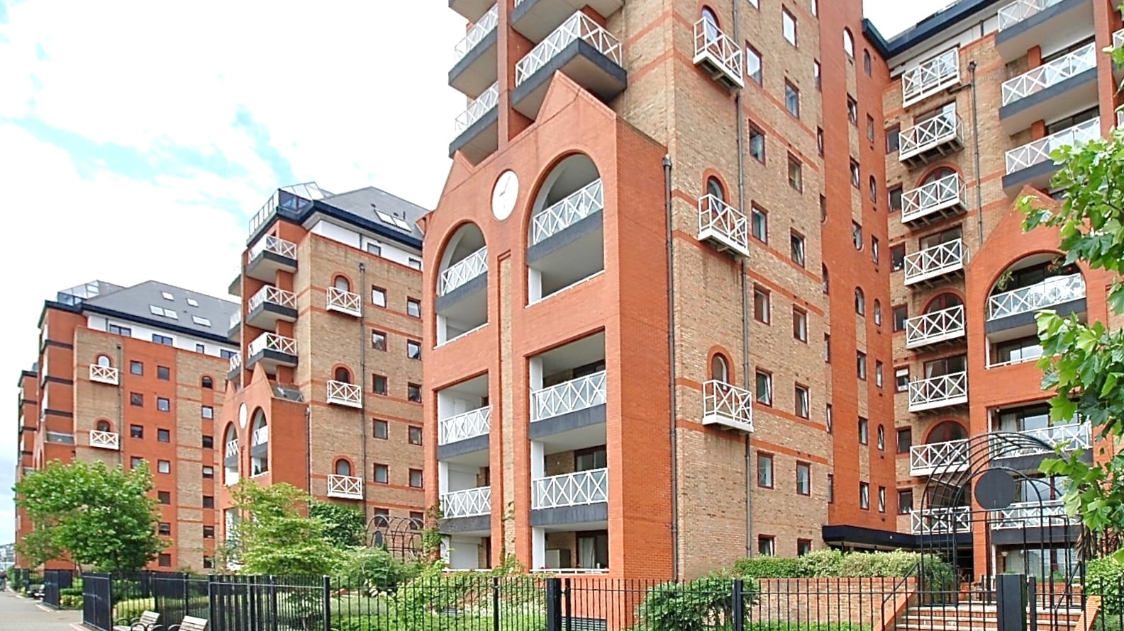Three of the brick blocks behind iron railings, with white lattice balconies and a clock set into an arched gable