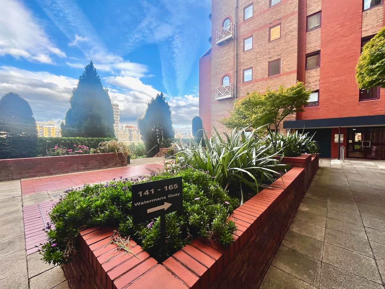 A brick planter with a sign reading 141 to 165 Watermans Quay, in a paved courtyard beside the building